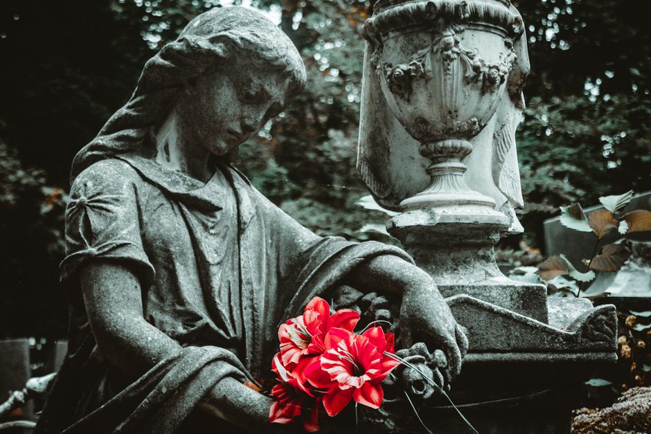 A weathered stone statue of a grieving woman with detailed facial features and flowing hair, dressed in a draped gown with puffed sleeves, holding a bouquet of vibrant red and white flowers with strip