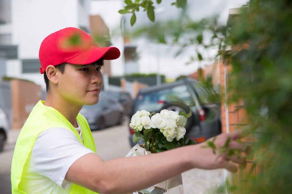 A young male flower delivery courier wearing a red cap and high-visibility yellow vest handing over a floral gift outside a residential building. The bouquet consists of white hydrangeas arranged in a